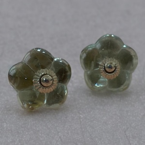 Puede incluir: Dos pomos de armario con forma de flor. Cada pomo tiene cinco pétalos de color verde translúcido. El centro de cada pomo es un círculo metálico dorado con un pequeño punto plateado en el centro. Los pomos son decorativos.