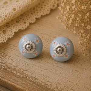 May include: Two light blue ceramic cabinet knobs with white and orange daisy patterns. Each knob has a silver-coloured metal centre and decorative trim. These decorative knobs are perfect for adding a touch of floral charm to furniture.