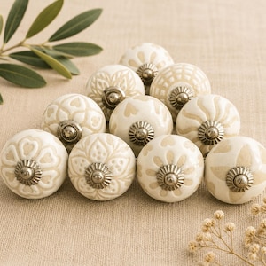 Può includere: Un gruppo di undici pomelli per mobili in ceramica bianco sporco con vari motivi floreali e geometrici in rilievo. Ogni pomello ha una base in metallo argentato. Disposti su una superficie di tessuto dai toni neutri.