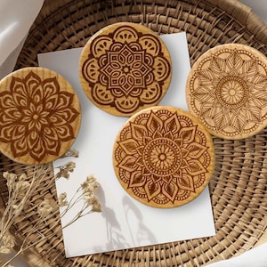 Può includere: Quattro sottobicchieri rotondi in legno con intricati disegni mandala. I sottobicchieri sono marrone chiaro con motivi incisi marrone scuro. Sono disposti su un vassoio di vimini intrecciato con un panno bianco e fiori secchi.