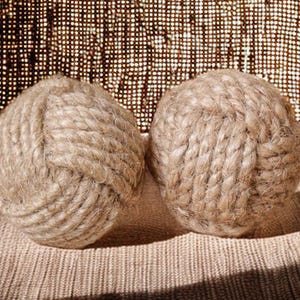 May include: Two beige rope balls, each intricately knotted. The balls are made of thick, natural-coloured rope, and sit on a textured, neutral-toned surface. The background features a woven fabric with a subtle pattern.