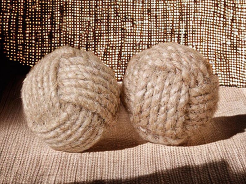 Pode incluir: Duas bolas de corda bege, cada uma intrincadamente amarrada. As bolas s&atilde;o feitas de corda grossa de cor natural e assentam sobre uma superf&iacute;cie texturizada de tom neutro. O fundo apresenta um tecido tecido com um padr&atilde;o sutil.