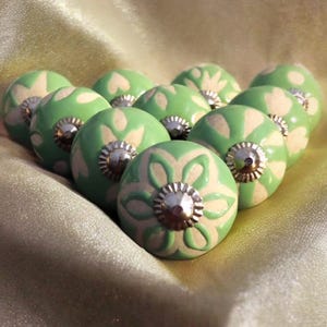 Może przedstawiać: Kolekcja zielonych i kremowych ceramicznych gałek do szafek z kwiatowymi wzorami. Każda gałka ma srebrną metalową podstawę. Gałki są ułożone na złotej tkaninie, podkreślając ich dekoracyjny charakter.