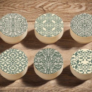 Può includere: Sei piccoli contenitori rotondi con coperchi decorativi. I coperchi presentano un motivo ripetuto in tonalità di verde e crema. I contenitori sono disposti su una superficie di legno.