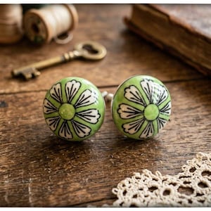 May include: A pair of green ceramic cabinet knobs with a floral design in white and black. They sit on a wooden surface, with a vintage key, thread spools, and a book in the background. Decorative hardware.