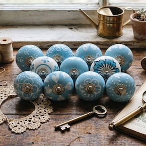 Op de afbeelding: Een verzameling blauwe en witte decoratieve kastknoppen met bloemen- en hartpatronen. De knoppen hebben goudkleurige metalen accenten. De afbeelding bevat ook een vintage sleutel, een vergrootglas en spoelen garen.