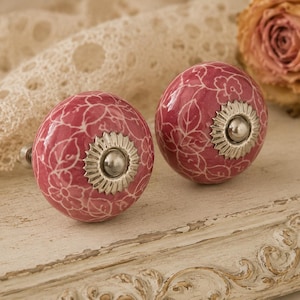 Puede incluir: Dos pomos de cerámica rosa con diseños florales blancos y herrajes plateados. Los pomos son redondos y tienen un estilo vintage decorativo. El fondo incluye tela de encaje y una rosa seca.