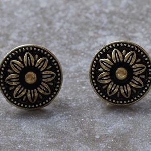 May include: A pair of round, antique gold-coloured stud earrings. Each earring has a detailed floral design in the centre, encircled by a dotted border. The earrings are set against a neutral, textured background.