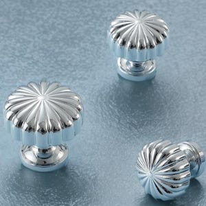 Puede incluir: Tres pomos de armario plateados con un diseño acanalado en forma de flor. Los pomos están colocados sobre un fondo azul claro texturizado. Estos pomos decorativos son adecuados para proyectos de mobiliario y decoración del hogar.