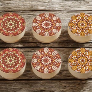 Peut inclure: Six petits contenants ronds avec des couvercles ornés d'un motif mandala détaillé dans des tons de rouge, orange et jaune. Les contenants ont une base beige et sont disposés sur une surface en bois.
