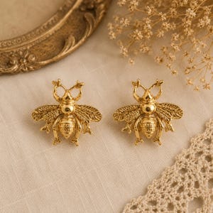 May include: A pair of gold-coloured bee stud earrings. The earrings feature detailed wings and body segments. The background includes a gold-framed mirror and dried flowers, suggesting a jewellery or accessory theme.