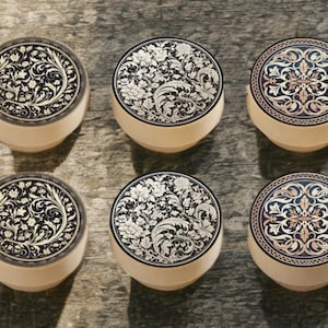 May include: Six decorative round boxes with ornate floral designs. The lids feature black, white, and gold patterns, while the bases are cream-coloured. The boxes are arranged on a textured, neutral-toned surface.