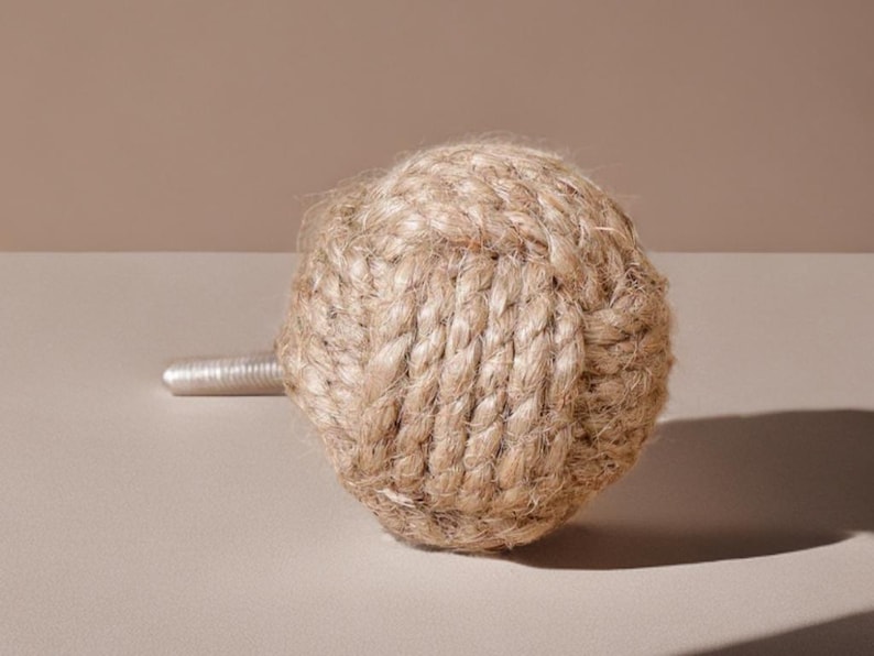 Pode incluir: Um puxador de porta decorativo com um design esf&eacute;rico feito de corda natural tran&ccedil;ada. O puxador tem uma haste de metal prateado para montagem. A bola de corda &eacute; de cor castanho claro, sobre um fundo neutro.