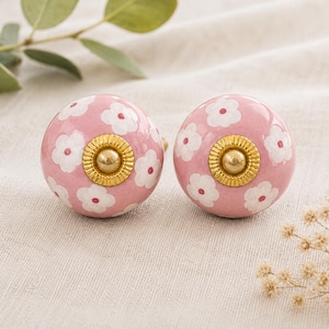 May include: Two pink ceramic cabinet knobs with white floral patterns and gold-colored metal accents. The knobs are round and feature a central gold-colored screw. Decorative hardware for drawers or cabinets.
