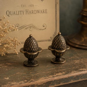 Puede incluir: Dos tiradores de armario antiguos en forma de bellota de latón. Los tiradores tienen un diseño detallado y texturizado y se asientan sobre una base decorativa. El fondo incluye un letrero vintage que dice "QUALITY HARDWARE" y el año "EST. 1924".