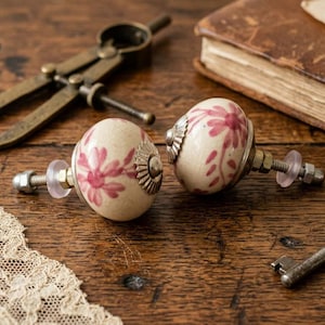 Puede incluir: Dos pomos de cerámica decorativos con un diseño floral en rosa y blanco, sobre una superficie de madera. Otros elementos incluyen una llave antigua, un compás de latón y un libro encuadernado en cuero.