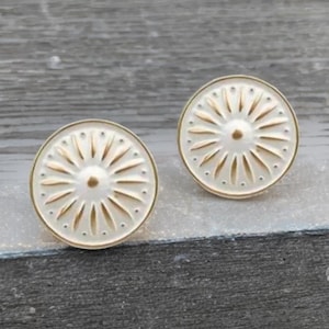 May include: Two circular earrings in off-white with a gold-coloured border and a sunburst pattern. Each earring features a central gold dot and radiating lines. The earrings have a vintage-inspired design. The background is a neutral grey.