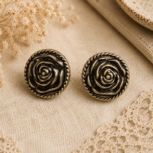May include: A pair of antique brass-coloured rose-shaped buttons. Each button has a detailed rose design within a twisted rope border. The buttons are set on a beige fabric, with dried flowers and lace trim in the background.