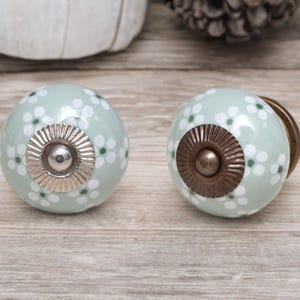 Op de afbeelding: Twee ronde, lichtgroene keramische kastknoppen met witte bloempatronen. De knoppen hebben zilveren en bronzen hardware. De knoppen liggen op een licht houten oppervlak.