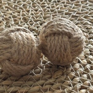 Pode incluir: Duas bolas decorativas de corda, cada uma meticulosamente trabalhada com fibra natural bem enrolada. As bolas s&atilde;o esf&eacute;ricas, com n&oacute;s vis&iacute;veis e uma superf&iacute;cie texturizada. Elas repousam sobre um fundo tecido castanho claro, sugerindo um tema n&aacute;utico ou r&uacute;stico.