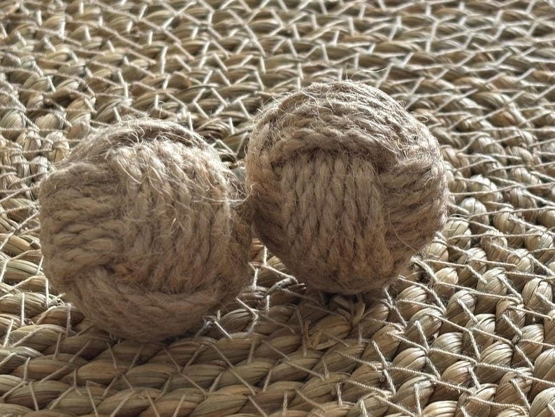 Pode incluir: Duas bolas decorativas de corda, cada uma meticulosamente trabalhada com fibra natural bem enrolada. As bolas s&atilde;o esf&eacute;ricas, com n&oacute;s vis&iacute;veis e uma superf&iacute;cie texturizada. Elas repousam sobre um fundo tecido castanho claro, sugerindo um tema n&aacute;utico ou r&uacute;stico.