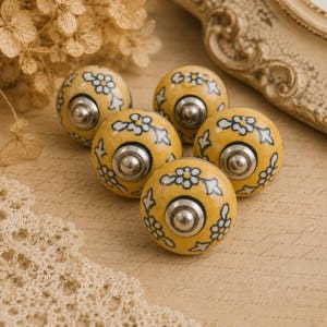 Könnte beinhalten: Fünf gelbe Keramik-Schrankknöpfe mit einem Blumenmuster in Weiß und Schwarz. Jeder Knopf hat einen silberfarbenen Metallsockel. Die Knöpfe sind auf einem Hintergrund im Vintage-Stil angeordnet.