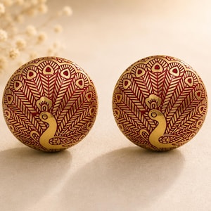 Op de afbeelding: Twee ronde, rood-gouden oorbellen met een pauwenontwerp. De oorbellen hebben een gouden pauw en gouden veerdetaillering. De achtergrond is neutraal.