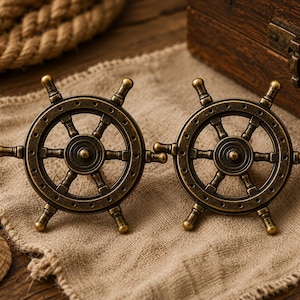 Puede incluir: Dos adornos decorativos de rueda de barco de latón antiguo. Cada rueda tiene seis radios y cuatro manijas. Las ruedas están sobre una tela beige con un cofre de madera y una cuerda en el fondo. También se ve una concha.