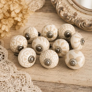 Può includere: Una collezione di pomelli per mobili in ceramica color crema con intricati motivi floreali e geometrici. Ogni pomello presenta una base e una vite in metallo argentato. I pomelli sono disposti su una superficie di carta in stile vintage, suggerendo un tema di arredamento per la casa.