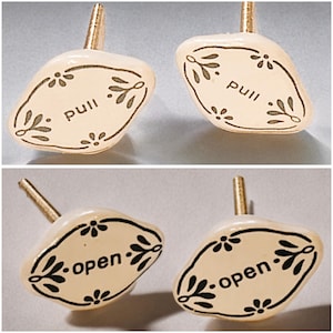 May include: Cream-coloured cabinet knobs with gold stems. The knobs have an oval shape with a black border and floral accents. The word "pull" is printed on two knobs, and "open" is printed on the other two.