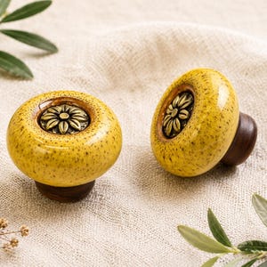 May include: Two yellow ceramic cabinet knobs with a speckled finish and a bronze flower design in the center. The knobs have a dark brown wooden base, perfect for adding a touch of elegance to furniture.