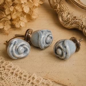Puede incluir: Tres pomos de armario de cerámica azul claro con forma de rosa y herrajes plateados. Los pomos están colocados sobre un fondo de papel vintage con flores secas y un espejo decorativo. Cada pomo tiene un tornillo para la instalación.