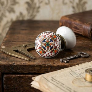 May include: Two decorative ceramic cabinet knobs with a floral design in red, orange, blue, and green on a white background. A vintage key, a brass tool, and a spool of thread are also on the wooden surface.