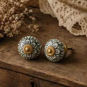 May include: Two ceramic cabinet knobs with a floral design in white and dark green. Each knob has a gold-coloured centre and a brass screw. The knobs are displayed on a wooden surface.