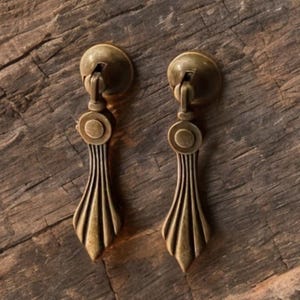 May include: A pair of antique brass drawer pulls with a decorative fan-shaped design. Each pull features a round backplate and a teardrop-shaped handle with vertical grooves. They are set against a rustic wooden background.