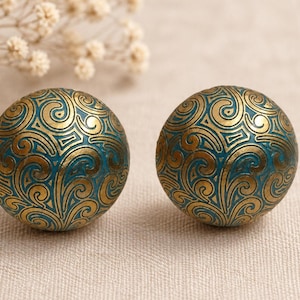 May include: Two spherical decorative objects with a teal base and gold swirling patterns. The objects have a metallic sheen. The background is a neutral beige, with a few sprigs of dried flowers.