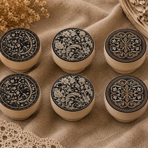 May include: Six decorative round boxes with floral designs. The lids feature intricate black and cream-coloured patterns. The boxes have a cream-coloured base and are arranged on a tan fabric surface.
