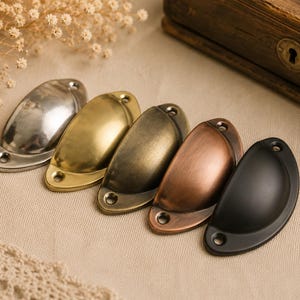 Könnte beinhalten: Fünf Metall-Muschelgriffe in verschiedenen Ausführungen: Silber, Messing, Bronze, Kupfer und Schwarz. Jeder Griff hat zwei Schraubenlöcher. Diese dekorativen Griffe sind ideal für Schränke und Schubladen.