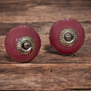 May include: Two round, burgundy cabinet knobs with silver and gold floral detailing. The knobs are set against a rustic wooden background, highlighting their decorative design. These knobs are ideal for adding a touch of elegance to furniture.