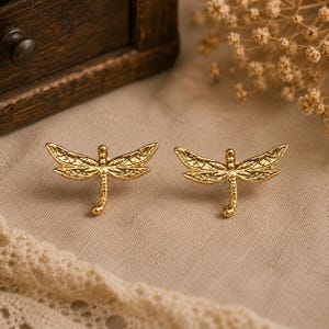 Peut inclure: Deux boucles d'oreilles dorées en forme de libellule. Chaque boucle d'oreille présente des ailes détaillées et un corps fin. Les boucles d'oreilles sont présentées sur un tissu de couleur neutre, avec un tiroir en bois en arrière-plan. Ce sont des bijoux.