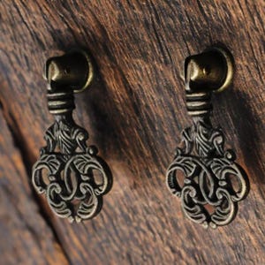 May include: Two antique bronze drawer pulls with ornate, scrollwork designs. Each pull has a decorative handle and a detailed, stylized base. The pulls are mounted on a textured, dark wood surface.