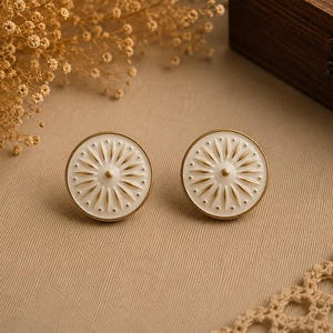Peut inclure: Une paire de boucles d'oreilles à clous blanches et dorées. Chaque boucle d'oreille présente un motif circulaire avec une base en émail blanc et des accents dorés. Le motif comprend un point central entouré de lignes rayonnantes et de petits points.