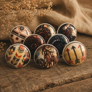 May include: Eight decorative cabinet knobs with diverse designs. The knobs display patterns such as arrows, geometric shapes, and floral motifs. The colour scheme includes cream, brown, red, and black, set against a wooden surface.