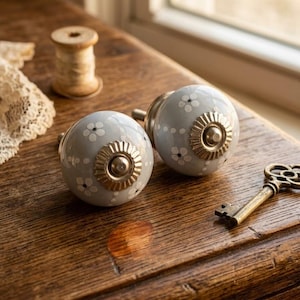Op de afbeelding: Twee lichtgrijze keramische kastknoppen met witte bloempatronen en messing accenten. Een vintage messing sleutel en een houten spoel liggen op een houten oppervlak. Een stuk kant is ook zichtbaar.