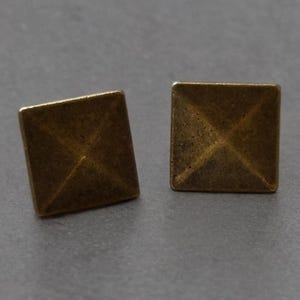 Pode incluir: Dois brincos quadrados, dourados. Cada brinco tem um design de pirâmide com uma superfície texturizada. Os brincos são exibidos em um fundo cinza. Estes brincos são um acessório simples, mas elegante.