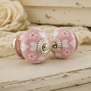 Puede incluir: Dos pomos de armario de cerámica rosa con diseños de corazones y flores blancos. Cada pomo tiene una base plateada y un centro decorativo plateado. Los pomos están colocados sobre un fondo de encaje y madera blanca desgastada.