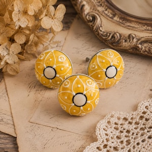 May include: Three yellow ceramic cabinet knobs with white floral designs and black accents. Each knob has a white centre. The knobs are on a vintage paper background, suggesting a decorative home decor item.