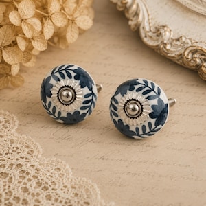 Puede incluir: Un par de gemelos de cerámica con base blanca y diseño floral azul. Cada gemelo presenta un centro de metal plateado y un pasador plateado. Los gemelos son redondos y tienen un patrón decorativo detallado.