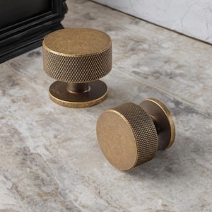 Op de afbeelding: Twee antieke messing kastknoppen. De knoppen hebben een getextureerd, cilindrisch lichaam en een cirkelvormige basis. De afwerking is een warme, verouderde messingkleur. Eén knop staat rechtop en de andere ligt op zijn kant.