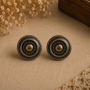 May include: A pair of antique bronze cabinet knobs. Each circular knob has a central raised circle, surrounded by dotted concentric rings and a rope-style edge. The knobs are displayed on a neutral beige surface.